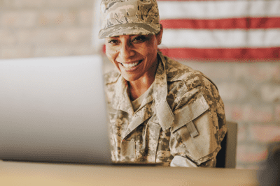 Smiling military woman in uniform at laptop with flag; NIKA's military per-diem furnished housing.
